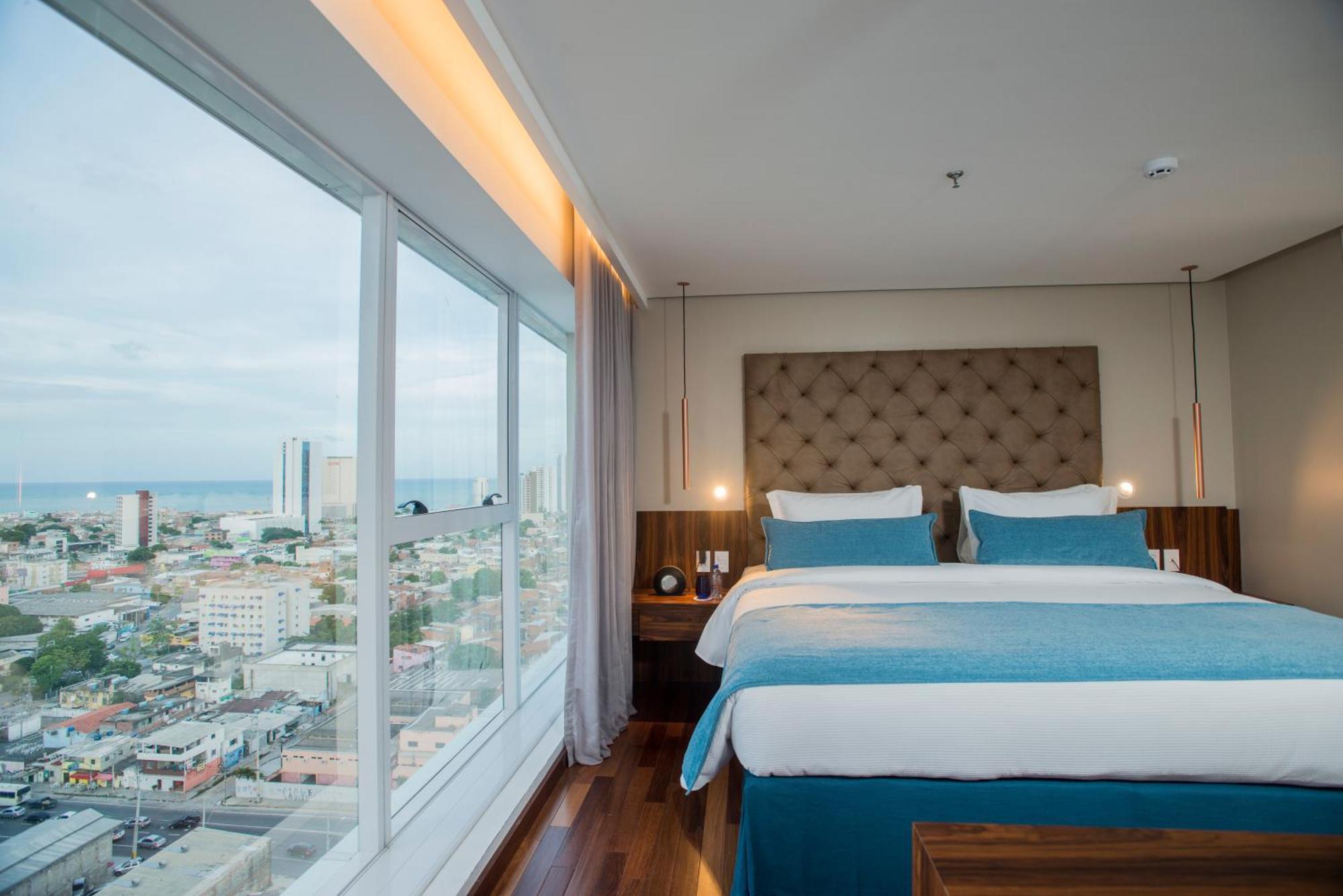 Hotel Luzeiros Recife Luaran gambar The photo shows a modern hotel room featuring a large window with a city view. Inside, there are two neatly made beds with light blue and white bedding, flanked by wooden nightstands. The room has a contemporary design, with a padded headboard and so